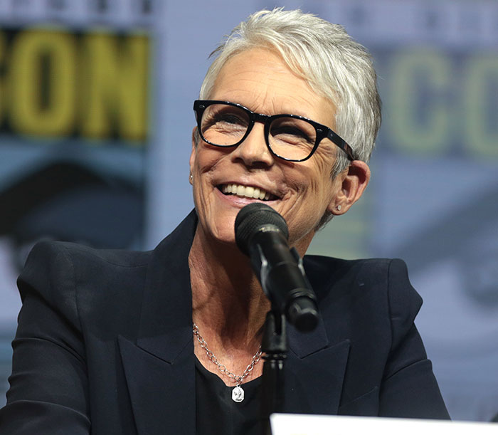 Jamie Lee Curtis speaking at an event, smiling and wearing glasses, related to plastic surgery discussion. Jamie Lee Curtis speaking at an event, smiling and wearing glasses, related to plastic surgery discussion.