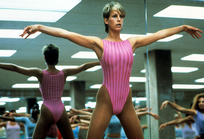 Jamie Lee Curtis in a pink leotard practicing dance moves in front of mirrors during a fitness class session. Jamie Lee Curtis in a pink leotard practicing dance moves in front of mirrors during a fitness class session.