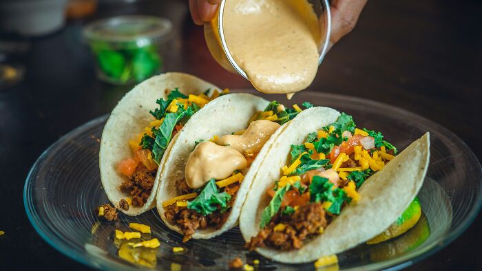 Three tacos filled with seasoned meat, kale, and cheese, with creamy sauce being poured on top, showcasing wild cooking opinions.
