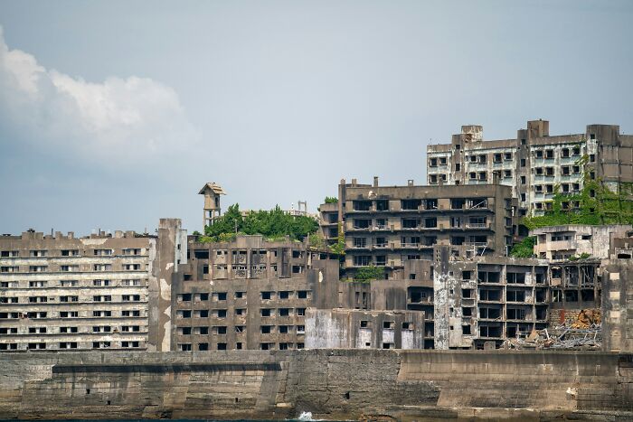 Abandoned tourist destination with decaying concrete buildings overgrown by vegetation near the water edge.