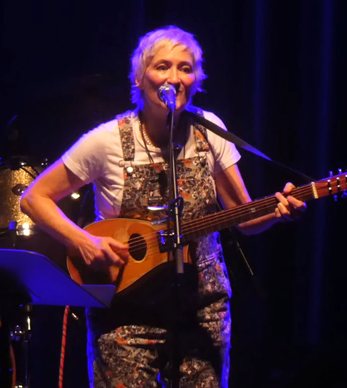 Singer Jill Sobule performing on stage with guitar, highlighted by stage lights during a live music event. Singer Jill Sobule performing on stage with guitar, highlighted by stage lights during a live music event.