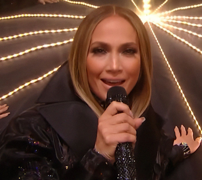 Jennifer Lopez holding a microphone, performing under bright lights at the AMAs during a controversial lesbian kiss moment.
