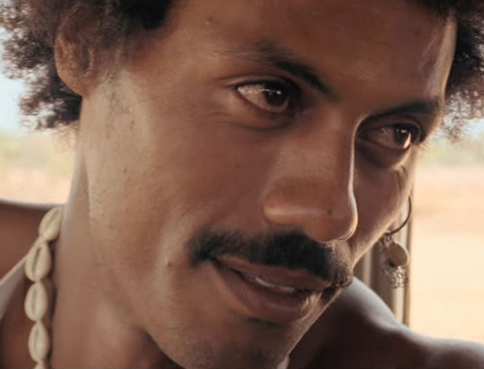 Close-up of a man with a mustache and curly hair wearing a shell necklace, highlighting natural facial features and expression.