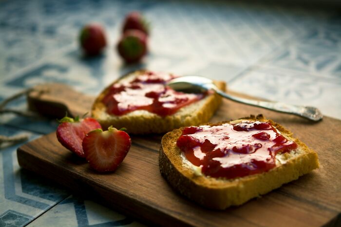 Two slices of bread with strawberry jam and fresh strawberries on a wooden board showing food prices comparison.