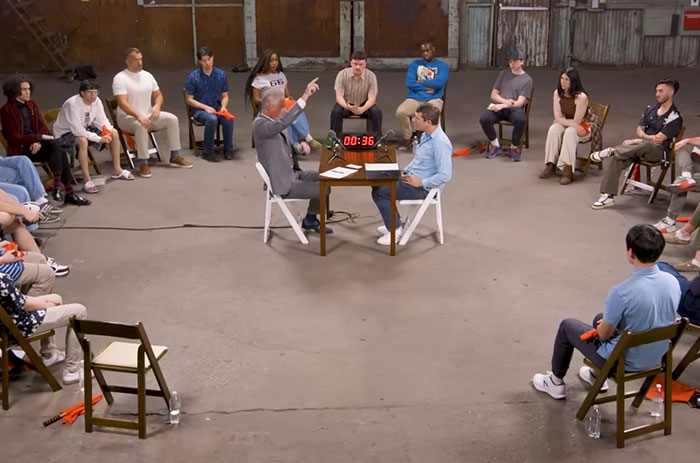 Jordan Peterson seated at a table debating atheists in a large room with an audience watching closely.
