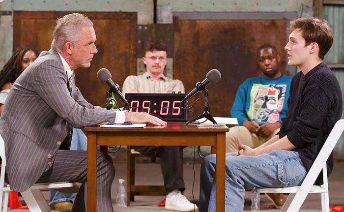 Jordan Peterson in a formal debate setting, facing a young atheist opponent, with microphones and a digital timer on the table.