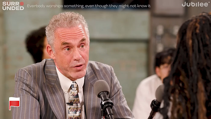 Jordan Peterson in a debate setting, wearing a striped suit, discussing ideas during a controversial atheist dialogue.