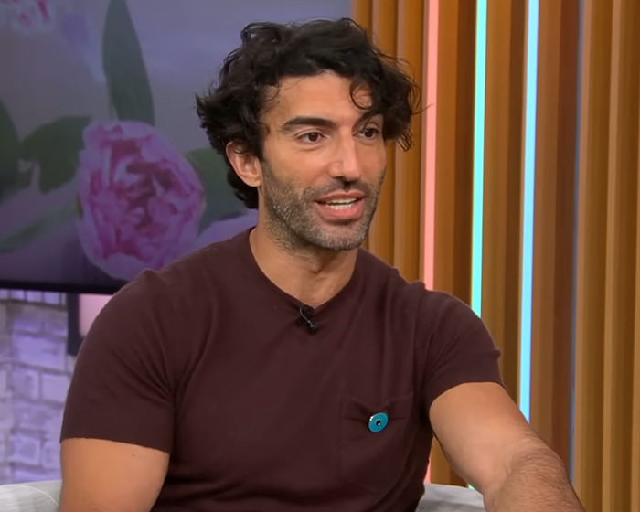 Man with curly dark hair and a beard wearing a brown shirt, speaking in a studio with colorful vertical lights behind him