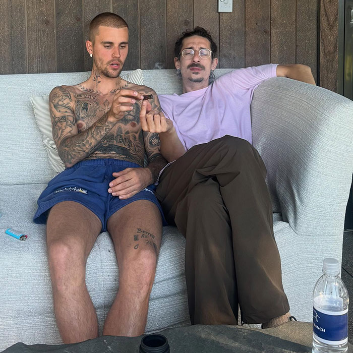 Justin Bieber shirtless with tattoos, holding smoking item, sitting next to man on couch in casual holiday setting.