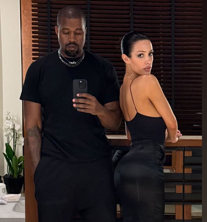 Bianca Censori posing in black outfit next to a man taking a mirror selfie in a room with wooden blinds.