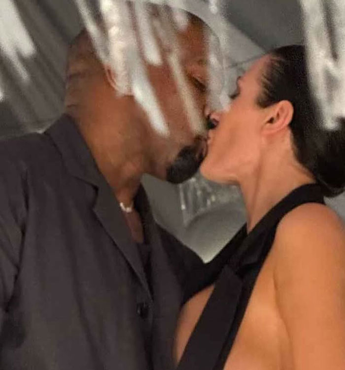 Bianca Censori in a revealing outfit kissing a man closely with a blurred background and soft lighting indoors.