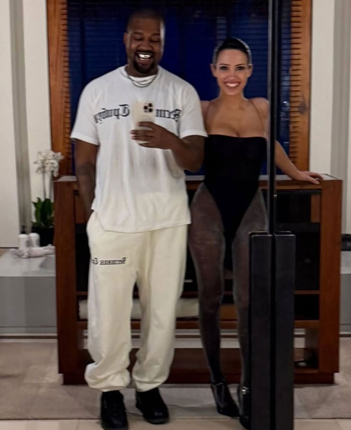 Kanye West's friend taking a mirror selfie with Bianca Censori wearing a revealing outfit in a modern indoor setting.