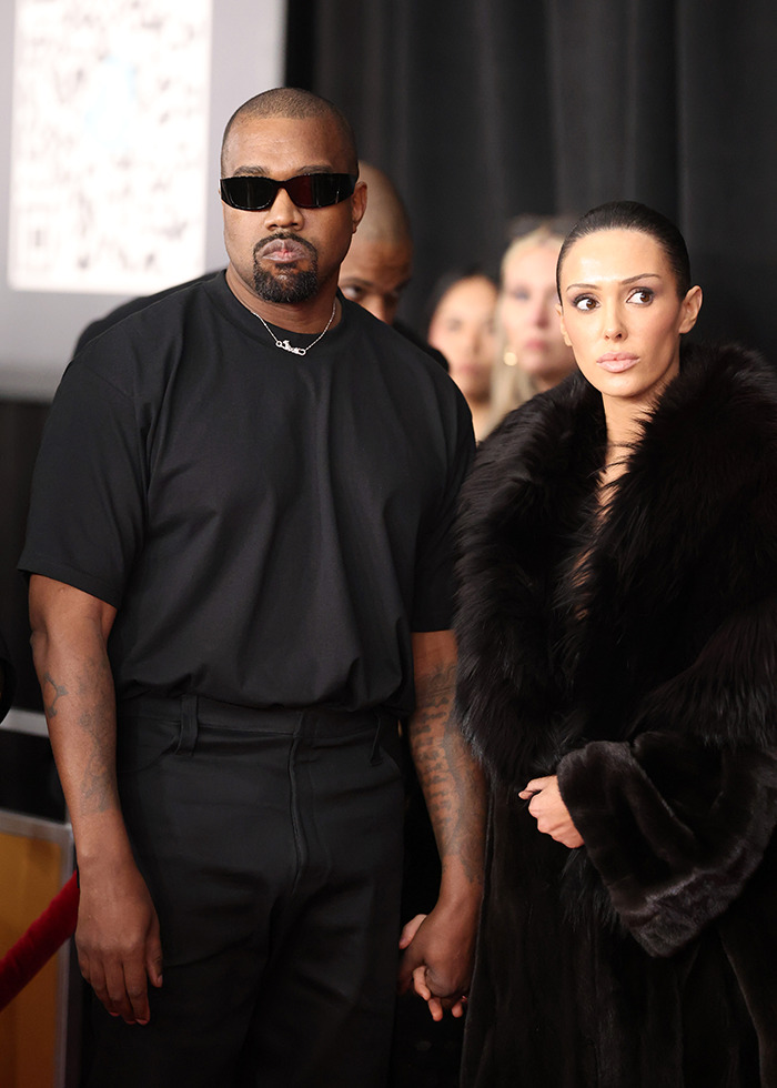 Kanye West wearing black and holding hands with Bianca Censori in a fur coat at an event.