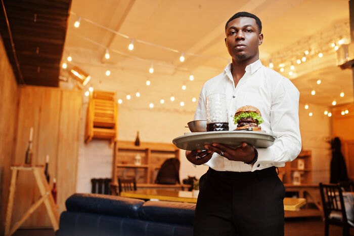 Young waiter serving burger and drink on tray in cozy restaurant, highlighting karen unhinged parents diabolical behavior context.