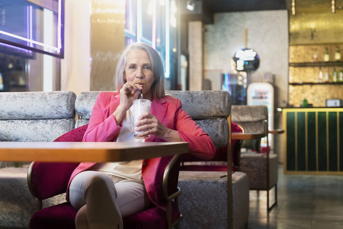 Older woman in pink blazer enjoying a drink at a cafe, reflecting the unhinged parents diabolical attitude.