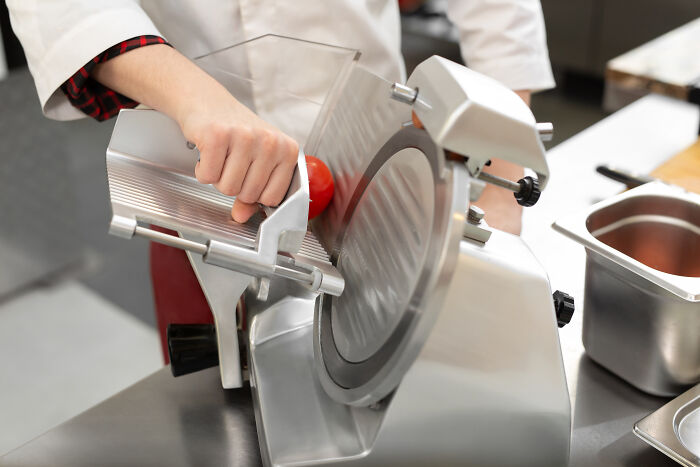 Person using a commercial slicer in a kitchen setting preparing food, highlighting precision with unhinged parents diabolical theme.