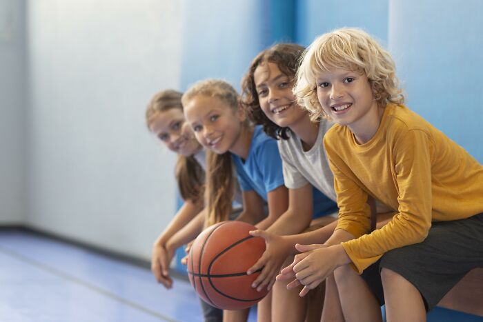Group of smiling kids sitting on gym bench with basketball, representing unhinged parents and diabolical behavior context