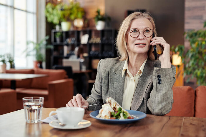 Middle-aged woman with glasses in a gray blazer on phone, appearing unhinged, in a cozy cafe setting with food and drink.