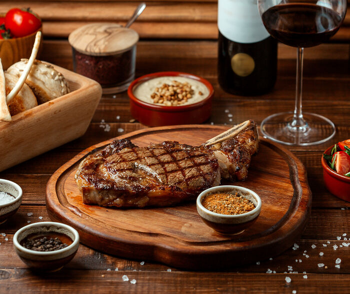 Grilled steak with seasonings on wooden board served alongside wine and fresh vegetables on rustic table setting.