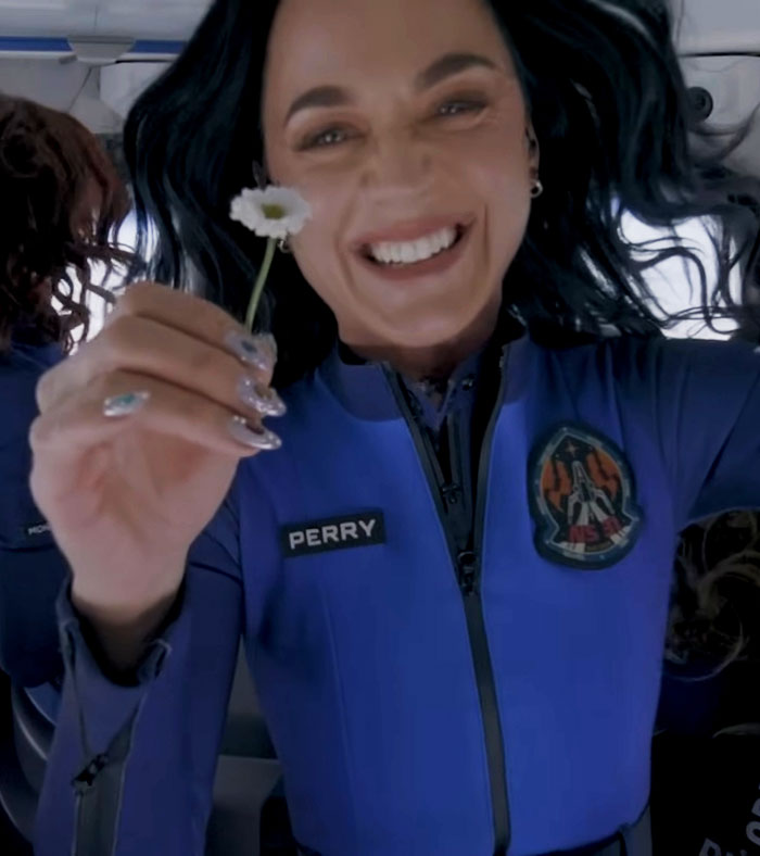 Katy Perry smiling and holding a small flower, wearing a blue suit with her name patch visible, close-up portrait.