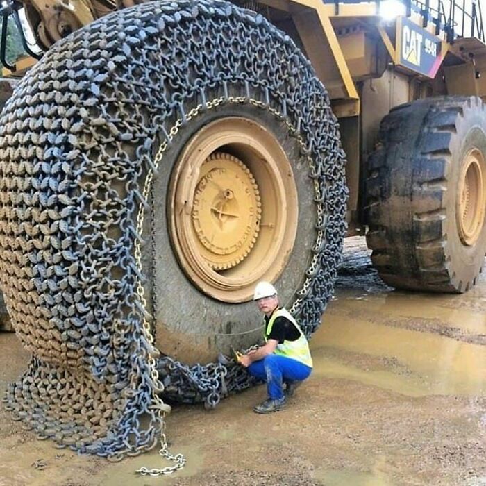 Large mining truck tire with heavy-duty chains and worker in safety gear adjusting chains on muddy ground, fascinating pictures.