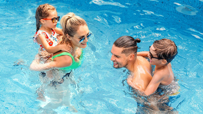 Family enjoying a sunny day at a condo pool, parents and kids playing and swimming together in clear blue water.