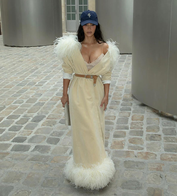 Kim Kardashian in a beige dress with feather details and a blue cap walking on cobblestone street during jewelry heist trial in Paris