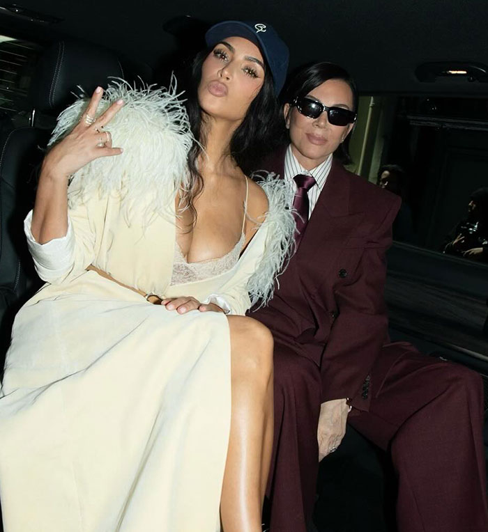 Kim Kardashian wearing a feathered outfit and cap, sitting next to a woman in sunglasses and a maroon suit.