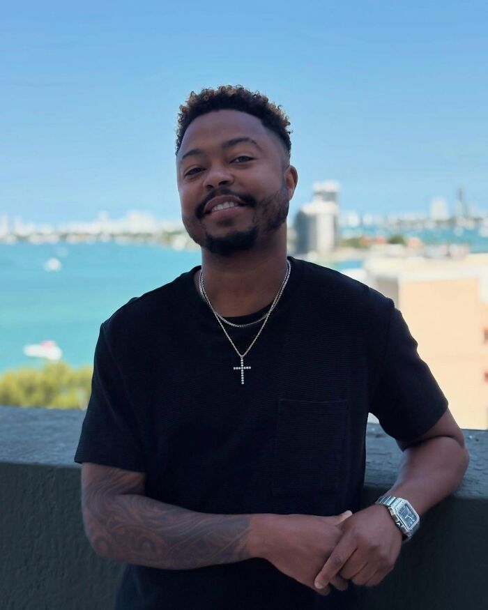Man smiling wearing black shirt and cross necklace with ocean and cityscape background notable people passed away in 2025.
