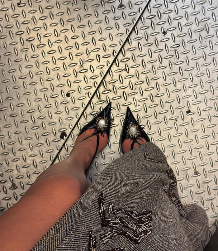 Close-up of black Met Gala stiletto heels with decorative embellishments on a diamond plate floor, showing a leg and patterned skirt. Close-up of black Met Gala stiletto heels with decorative embellishments on a diamond plate floor, showing a leg and patterned skirt.