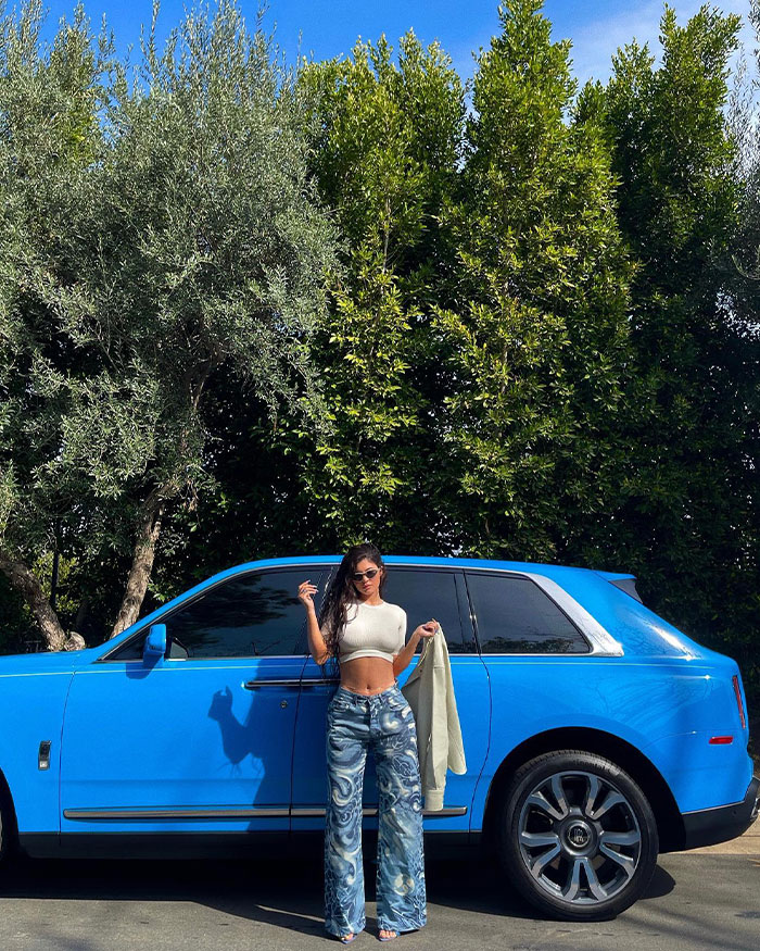 Kylie Junner posing by a blue luxury car surrounded by trees, highlighting Kylie Jenner&rsquo;s net worth and financial status update.