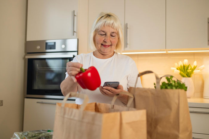 Woman holding red bell pepper and phone while unpacking groceries in kitchen, related to grocery Karen shopping dispute.