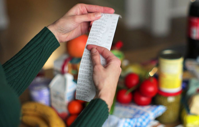 Person holding a grocery receipt with various fresh produce and packaged food items on a table nearby.