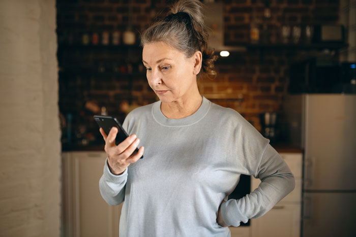 Middle-aged woman looking at smartphone with concern in kitchen, representing grocery Karen refusing to pay for extra items.