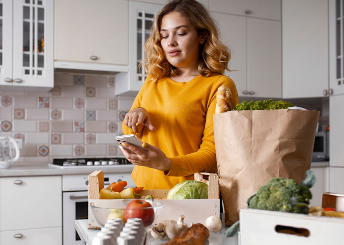 Woman in kitchen with groceries, using phone, illustrating grocery Karen refusing to pay for extra items scenario.