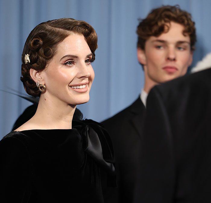 Lana Del Rey smiling with elaborate hairstyle at the Met Gala, with a young man in the background gaining internet attention.