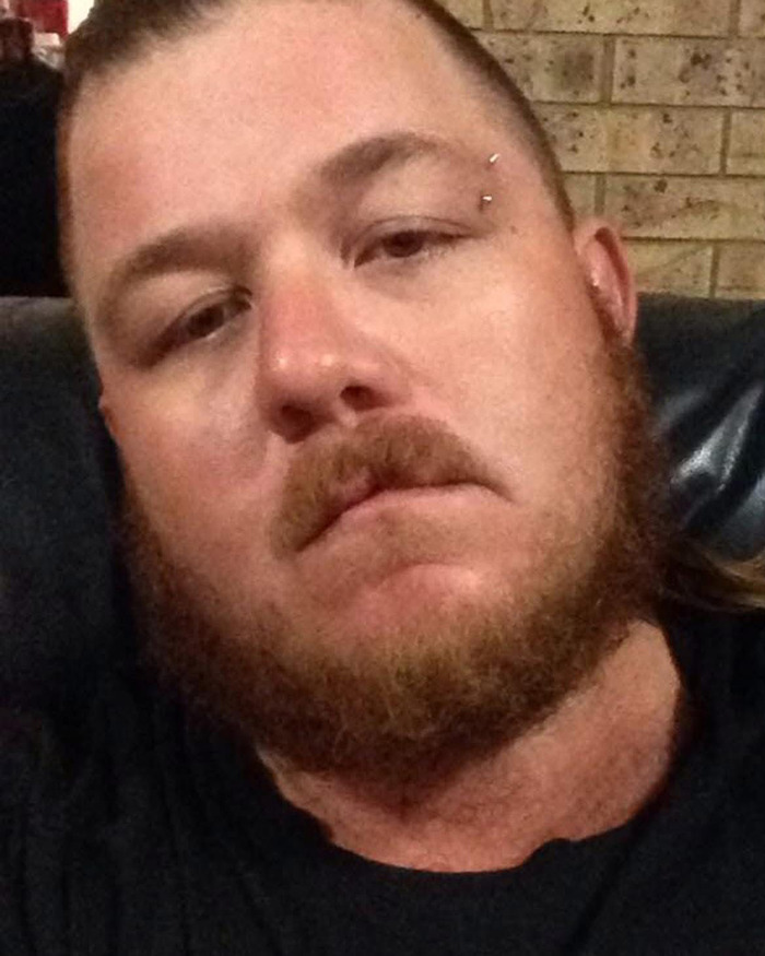 Man with a beard and eyebrow piercing sitting indoors against a brick wall background in a casual setting.