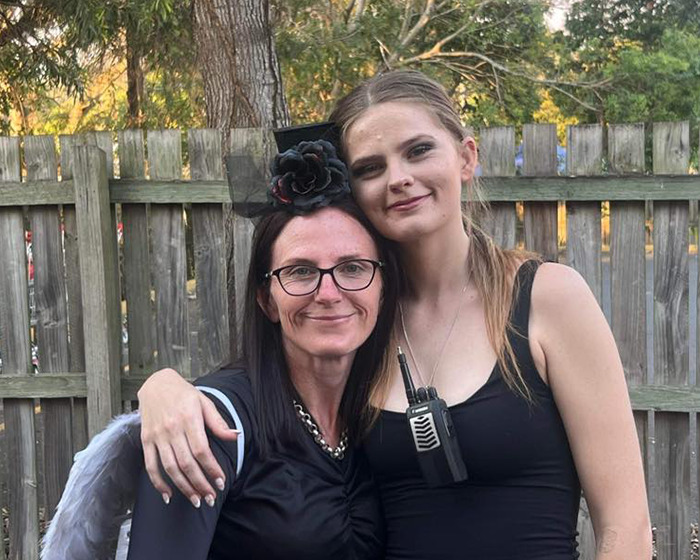 Two women posing together outside near a wooden fence, one wearing glasses and a black flower headpiece.
