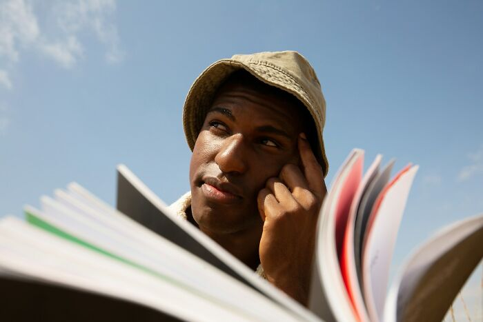 Man wearing a bucket hat thinking deeply while flipping through a large book under a clear blue sky, boomer opinions concept.