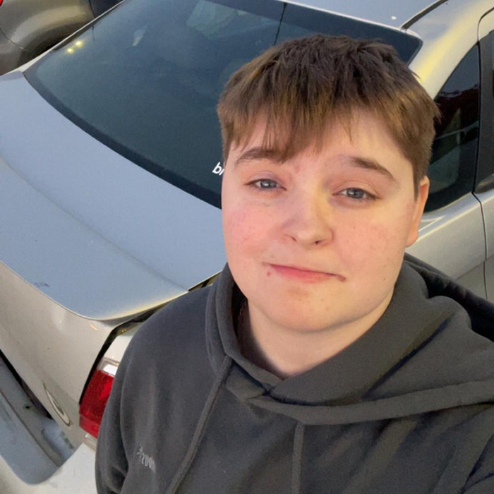 Person with short hair wearing a dark hoodie standing near a silver car, related to lesbian beaten unconscious McDonald's bathroom incident