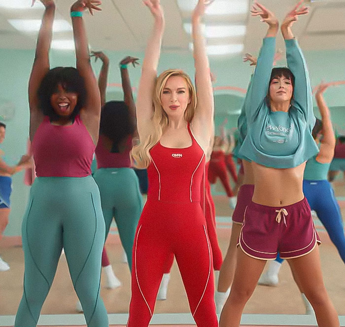 Lindsay Lohan leading a group in a fitness class, wearing a red outfit with no visible freckles in a bright studio.