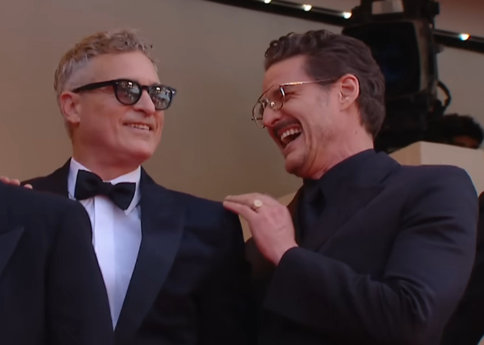 Pedro Pascal and Joaquin Phoenix smiling together while a lip reader decodes their conversation after an awkward hand wave.