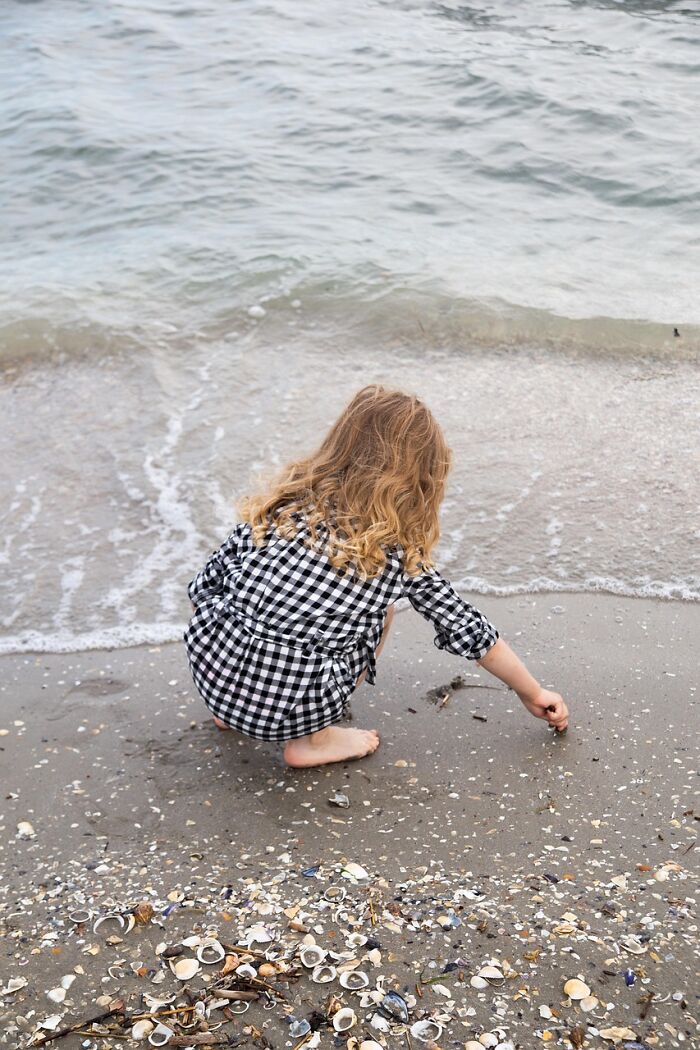 Young child with a hilariously bad name playing barefoot on a shell-covered beach near the water’s edge.