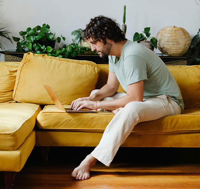 Person using the anonymity of the internet to share secrets while typing on a laptop seated on a yellow couch.