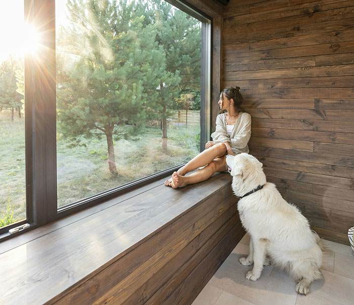 Woman sitting by a large window with a dog, reflecting in solitude, symbolizing people using internet anonymity.