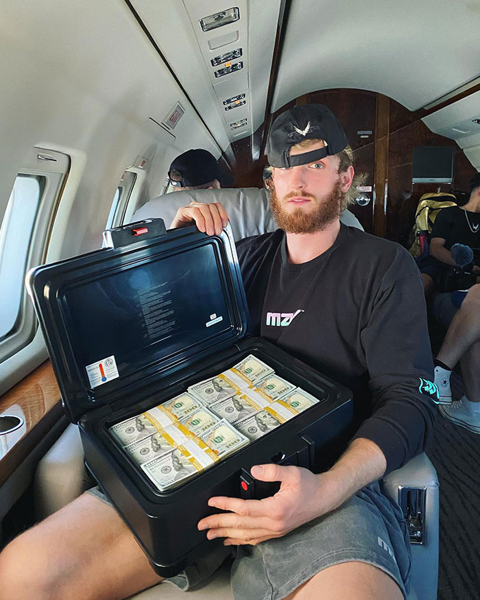 Logan Paul sitting in a private jet holding an open cash box filled with stacks of hundred-dollar bills, showcasing net worth.