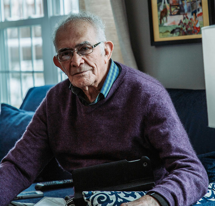Elderly man wearing glasses and a purple sweater sitting on a couch, portraying a long-lost grandfather concept.