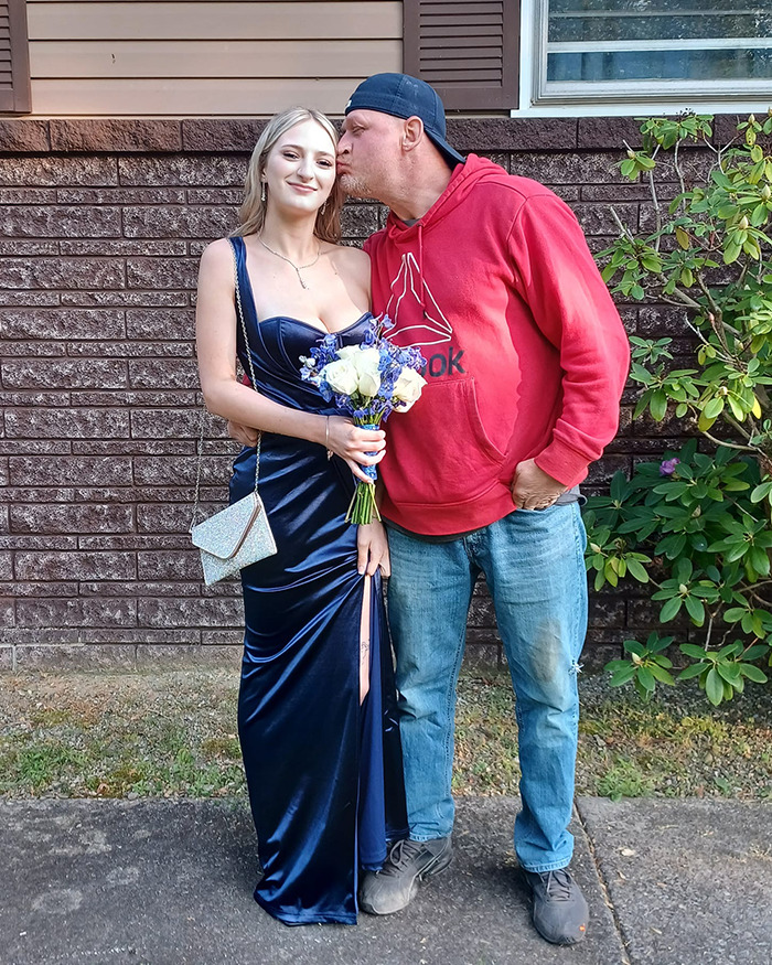Father and daughter posing outside a house, with the father kissing daughter's cheek, both dressed for a special occasion.