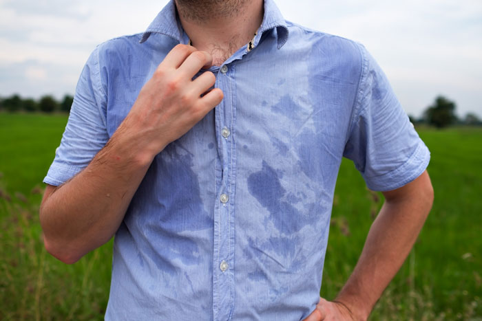 Man wearing blue shirt with large wet stains on the front, standing outdoors in a green garden area.