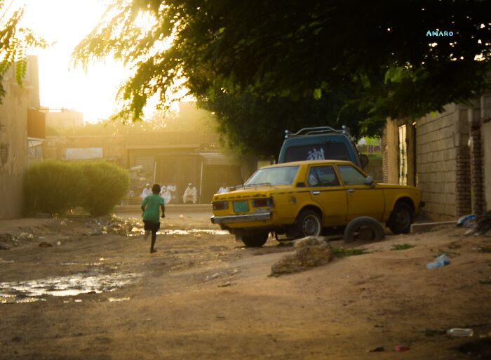 Child running on a dusty street near old yellow car under tree shade, depicting a major event not covered by mainstream media today.